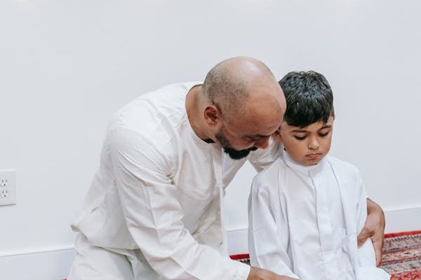 a-father-kneels-with-his-son-in-prayer-both-wearing-white-robes-against-a-backdrop-of-string-lights.-9127577-600x900